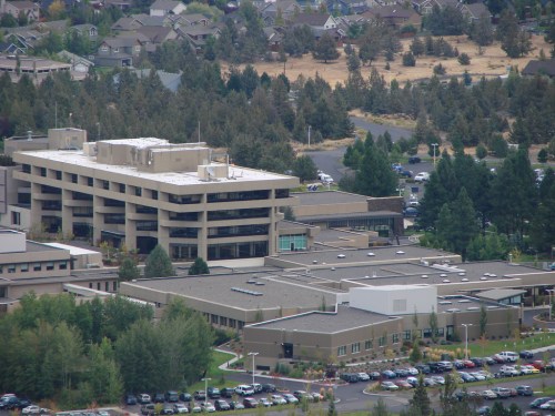 Aerial view of a large modern building surrounded by trees and parked cars, with a residential area in the background.