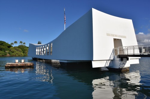 The USS Arizona Memorial, a white structure over water, with a flag and palm trees in the background under a blue sky.