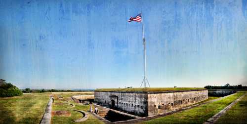 A historic fort with an American flag flying, surrounded by green grass and a clear blue sky. Visitors explore the area.