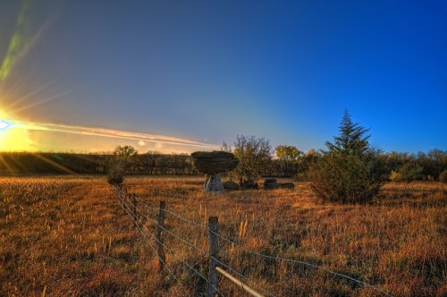 A serene landscape at sunset, featuring a grassy field, a wooden fence, and a large haystack under a clear blue sky.