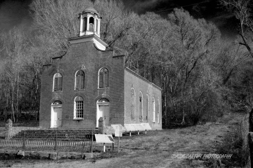 A historic brick church with a bell tower, surrounded by trees, captured in black and white.