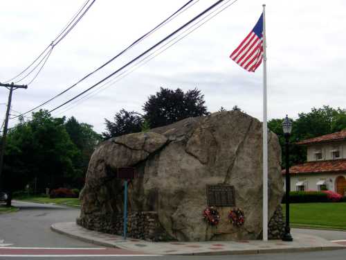 A large rock monument with a flagpole and American flag, surrounded by flowers and historical plaques.