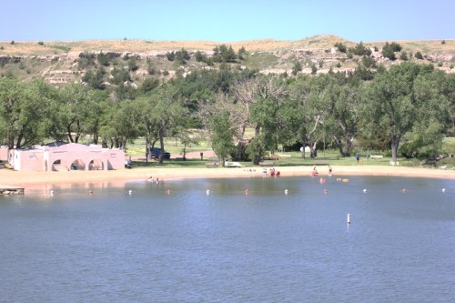 A serene lake scene with sandy beach, trees, and people enjoying the water on a sunny day.