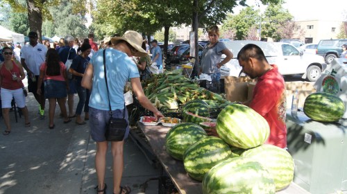 A bustling farmers market with people browsing fresh produce, including watermelons and corn, under sunny skies.