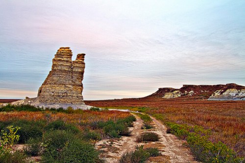 A rocky formation stands beside a dirt path, surrounded by colorful grasslands under a pastel sky.