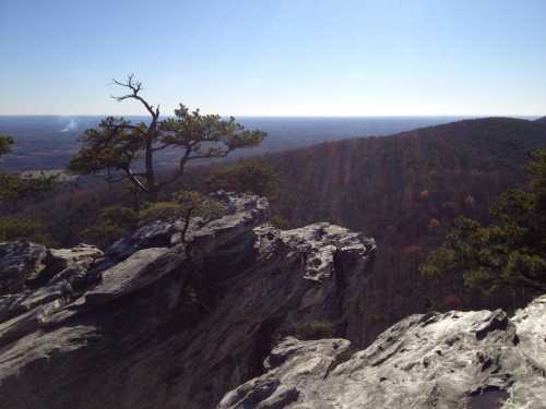 A rocky cliff overlooks a vast landscape of rolling hills and trees under a clear blue sky.