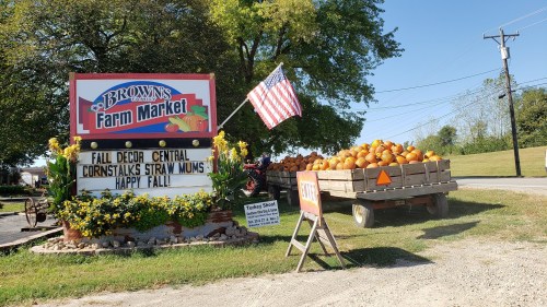 A farm market sign displays fall decor, cornstalks, and pumpkins, with a wagon full of pumpkins and an American flag nearby.