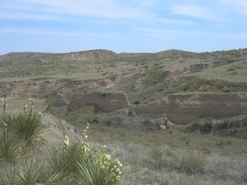 A scenic view of rolling hills and grassy terrain with a dry, eroded landscape under a blue sky.
