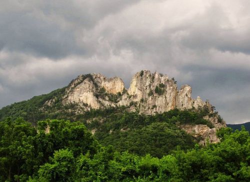 A rugged mountain peak rises above lush green trees under a cloudy sky.