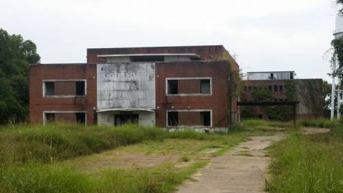 Abandoned brick building with overgrown grass and a cloudy sky in the background.