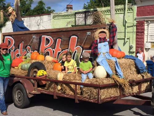 A festive float decorated with pumpkins, hay, and scarecrows, featuring two children sitting on bales of hay.