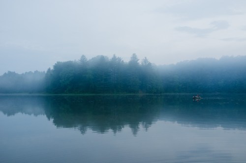 A misty lake scene with a forested island and a small boat on the calm water, reflecting the foggy surroundings.