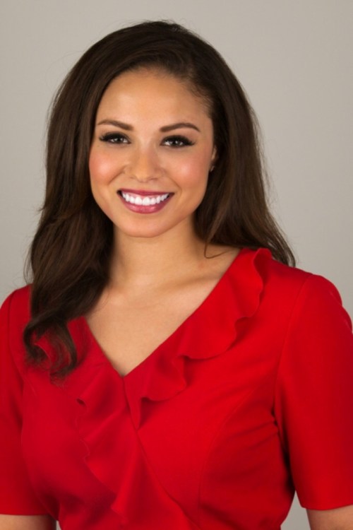 A smiling woman with long dark hair wearing a red blouse with ruffles, against a neutral background.