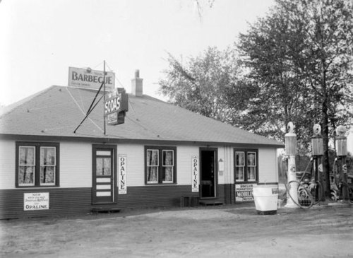 Historic barbecue restaurant with a sign, gas pumps, and trees in the background. Black and white photo.