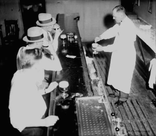 A bartender serves drinks to patrons at a vintage bar, with men in hats seated at the counter.