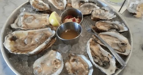A platter of fresh oysters on ice, served with lemon, cocktail sauce, and mignonette in small cups.