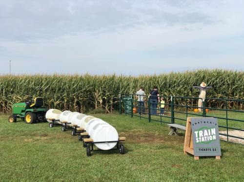 A train station with a green tractor and a line of white barrels, set against a cornfield and cloudy sky.