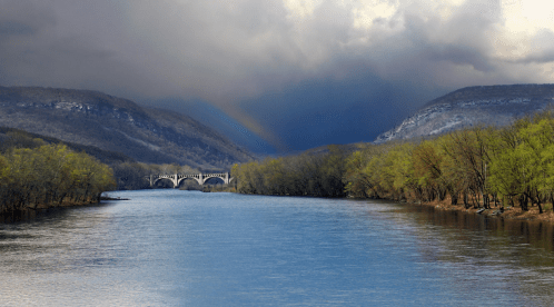A serene river flows between mountains, with a bridge in the distance under a cloudy sky. Green trees line the banks.