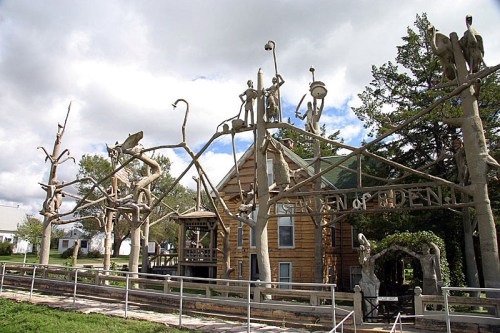 A wooden sculpture garden featuring intricate tree-like structures and figures, with a building in the background.
