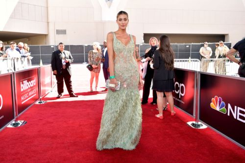 A woman in a shimmering green gown with fringe poses on the red carpet at an awards event.