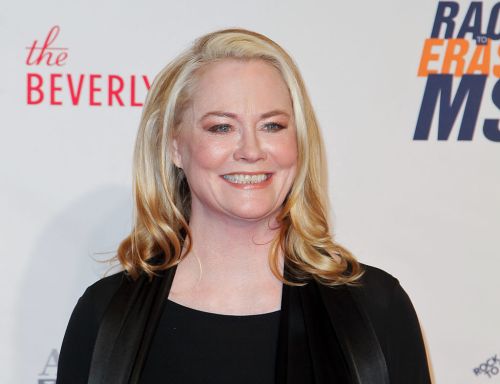 A smiling woman with long blonde hair poses at a red carpet event, wearing a black outfit.