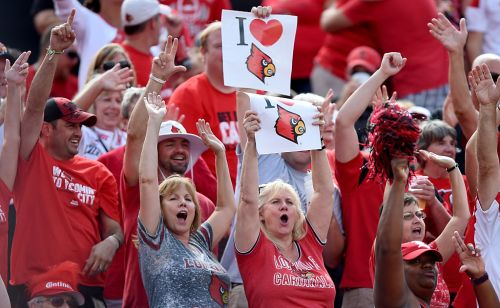 A crowd of enthusiastic fans in red shirts cheer, holding signs that say "I ❤️ Cardinals" at a sports event.
