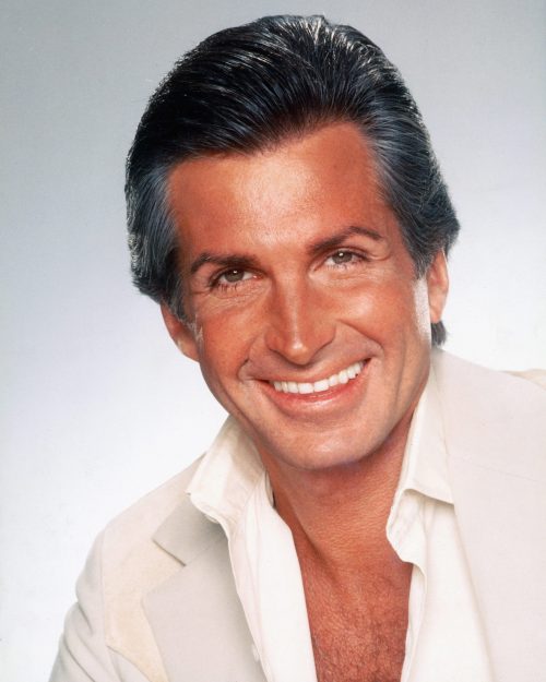 A smiling man with slicked-back hair, wearing a light-colored suit jacket over a white shirt, against a soft background.