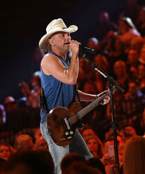 A musician in a cowboy hat sings into a microphone while playing guitar on stage, with a cheering crowd in the background.