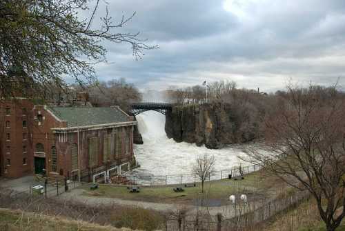 A waterfall cascades over rocky cliffs beside an old brick building, under a cloudy sky. Trees line the area.