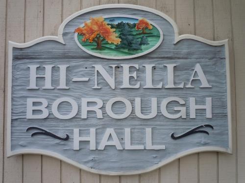 Sign for Hi-Nella Borough Hall featuring decorative trees and a scenic background.
