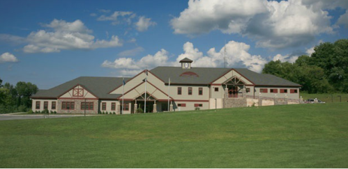 A large, modern building with a clock tower, set on a green lawn under a blue sky with fluffy clouds.