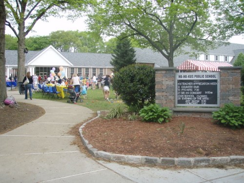 A school yard with tables, balloons, and people gathered for an event, featuring a sign for the school.