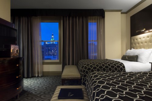 A hotel room with two beds, a bench, and a window showing a city skyline at dusk.