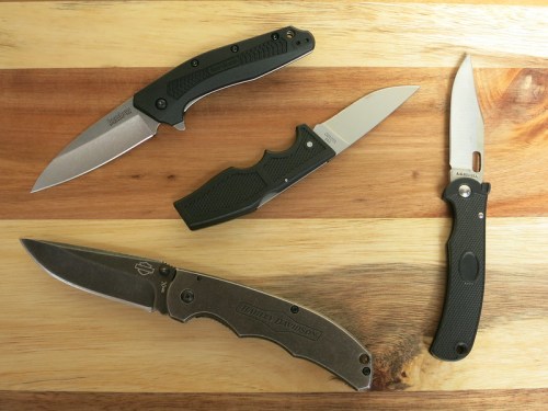 Four different folding knives displayed on a wooden surface, showcasing various blade shapes and handle designs.