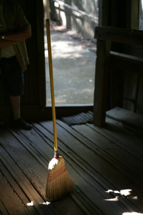 A broom stands upright on wooden floorboards, illuminated by sunlight streaming through an open doorway.
