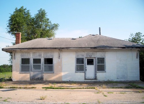 Abandoned single-story building with a weathered exterior, broken windows, and a chimney, set against a clear blue sky.