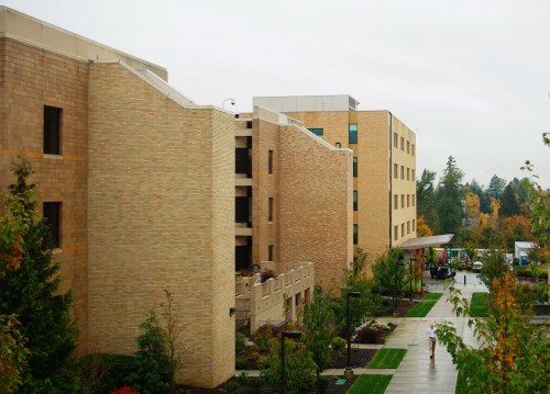 A view of modern brick buildings on a campus, surrounded by greenery and a cloudy sky.