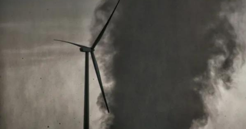 A wind turbine stands beside a swirling column of dark smoke against a cloudy sky.