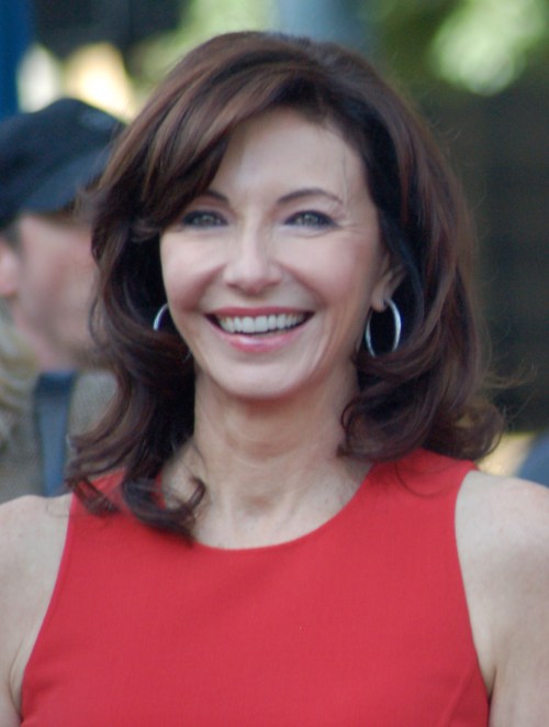 A woman with long, wavy brown hair smiles warmly, wearing a red dress and hoop earrings.
