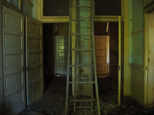 An old wooden ladder stands in a dimly lit room with multiple doorways and peeling paint on the walls.