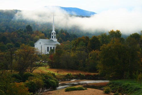 A white church with a tall steeple surrounded by trees and misty mountains, reflecting a serene autumn landscape.