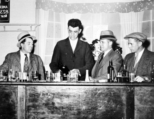 Four men in vintage attire stand at a bar, with one pouring a drink, surrounded by glasses and bottles.