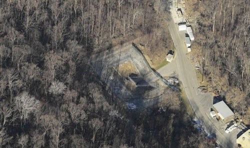Aerial view of a wooded area with a clearing, road, and several structures visible in the landscape.