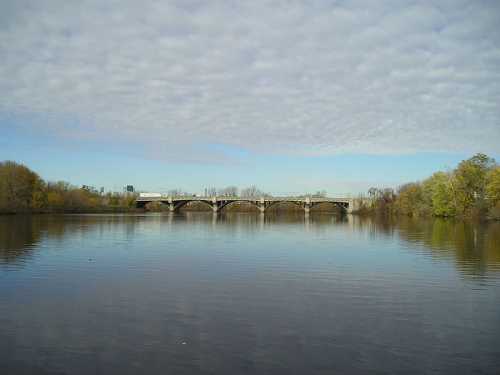 A calm river reflects a cloudy sky, with a bridge spanning across the water and trees lining the banks.