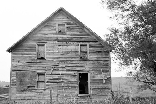 A weathered, abandoned wooden house with broken windows and a door ajar, surrounded by overgrown grass.