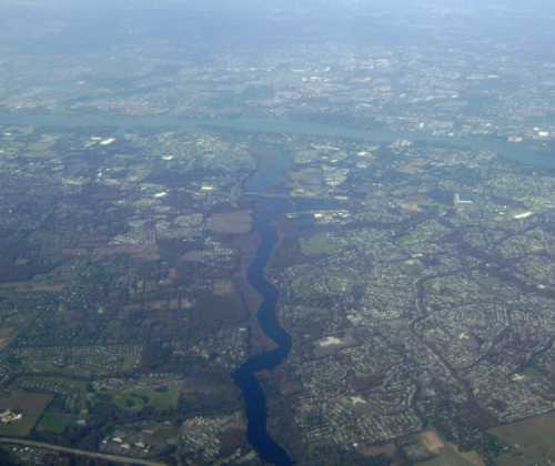 Aerial view of a river winding through a landscape of fields and suburban areas under a hazy sky.