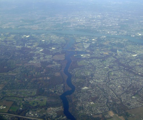Aerial view of a river winding through a landscape of fields and suburban areas under a hazy sky.