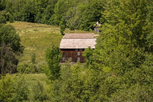 A rustic wooden barn nestled among lush green trees and rolling hills under a clear blue sky.