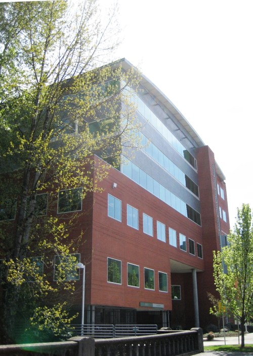 Modern multi-story building with large windows, surrounded by trees and bright sunlight.