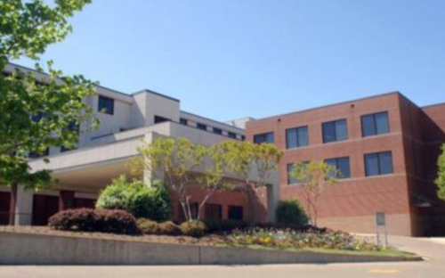 A modern building with a landscaped entrance, featuring trees and shrubs under a clear blue sky.